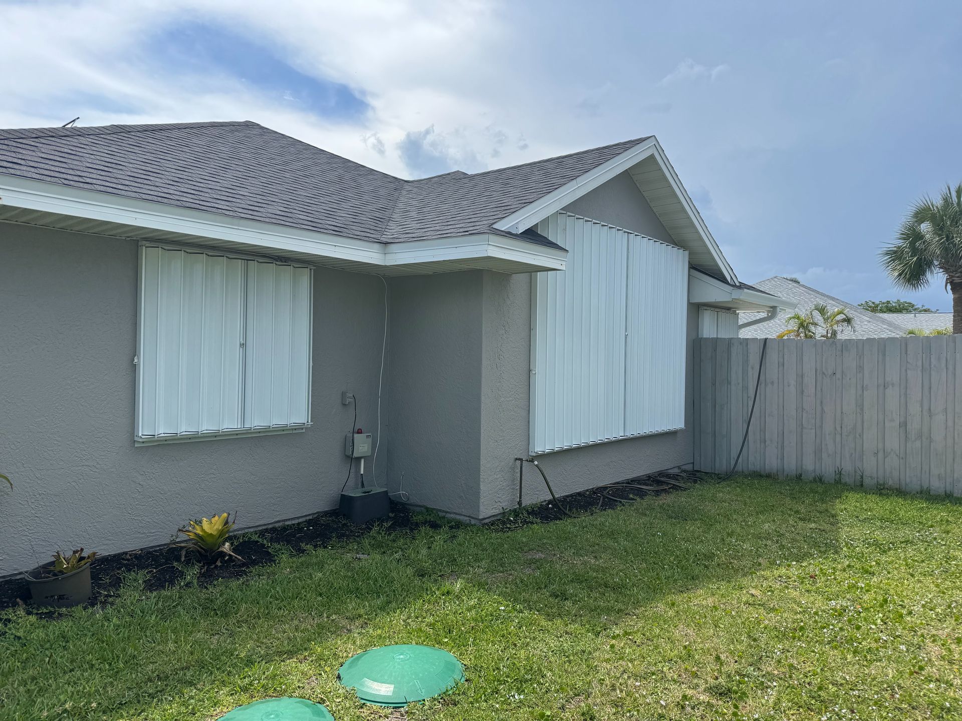 Side view of a gray house with white hurricane shutters closed over two windows, set in a grassy yard under a blue sky.