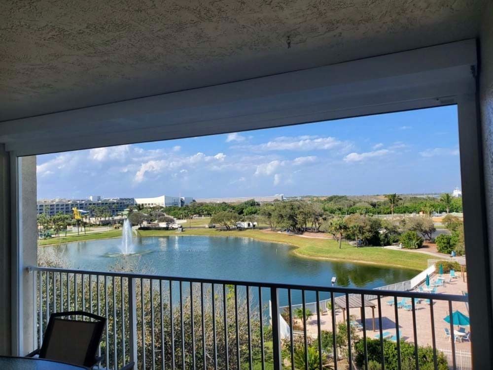 View from a balcony overlooking a pond with a fountain, green trees, and a pool deck under a bright blue sky.