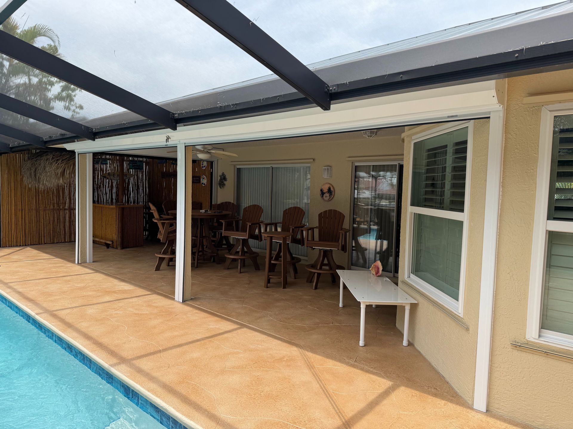 Covered outdoor pool patio with a tan textured deck, dining table with high chairs, and a white side table.