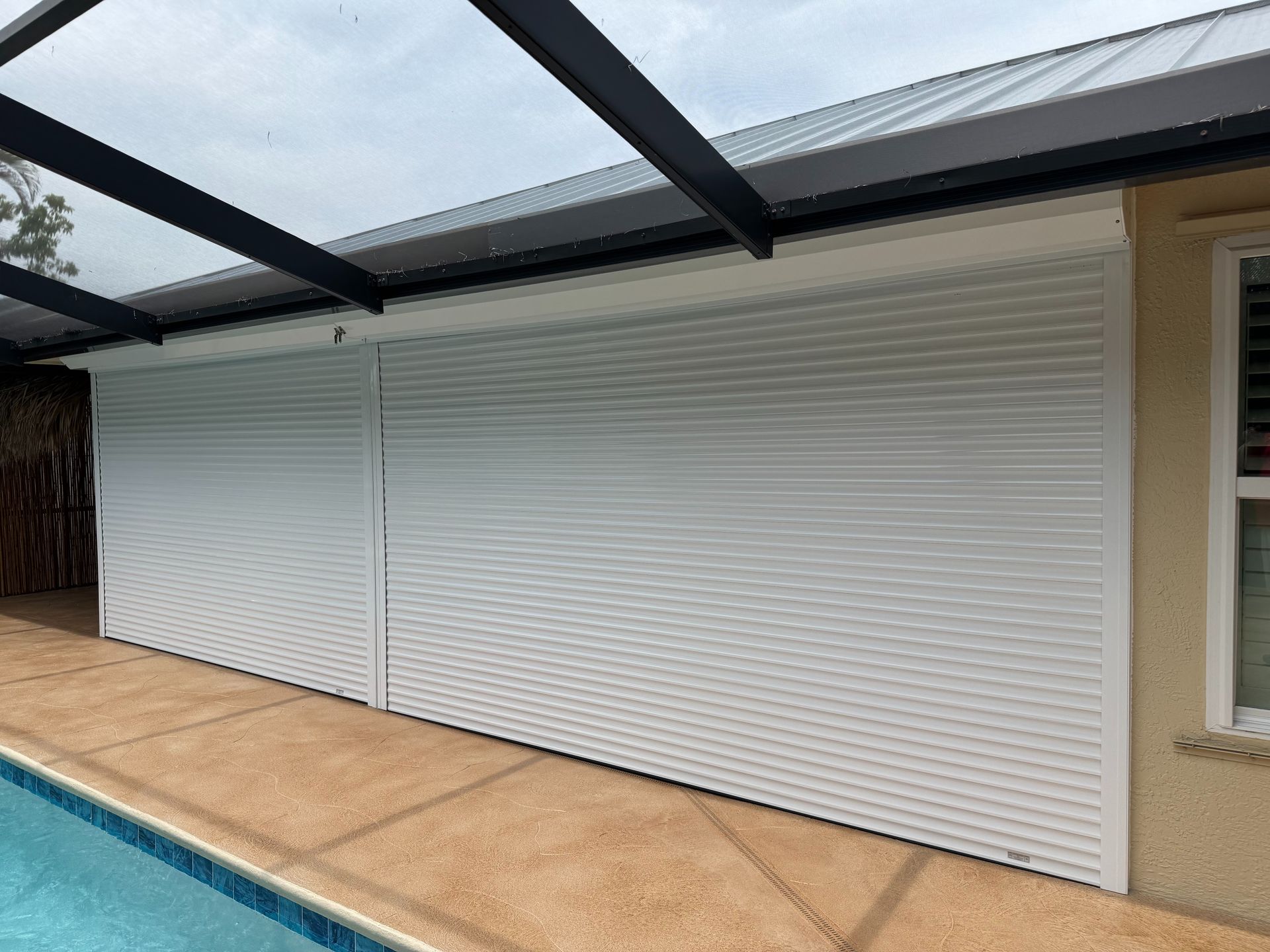 White accordion hurricane shutters are fully closed across a patio, protecting a home next to a swimming pool.