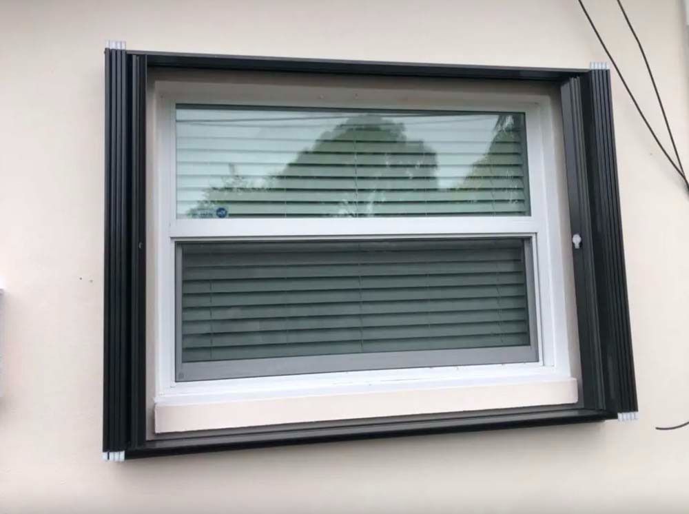A white-framed window with horizontal blinds, mounted on a light-colored wall, flanked by black accordion storm shutters.