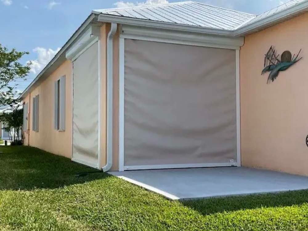 A tan house with closed retractable hurricane shutters over the garage and side window, set against a bright blue sky.