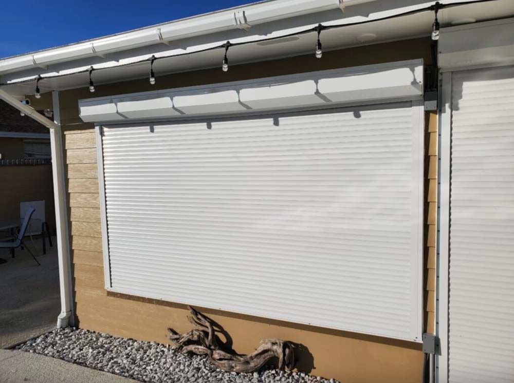 White horizontal security shutters installed over an exterior house window, mounted on light brown siding.