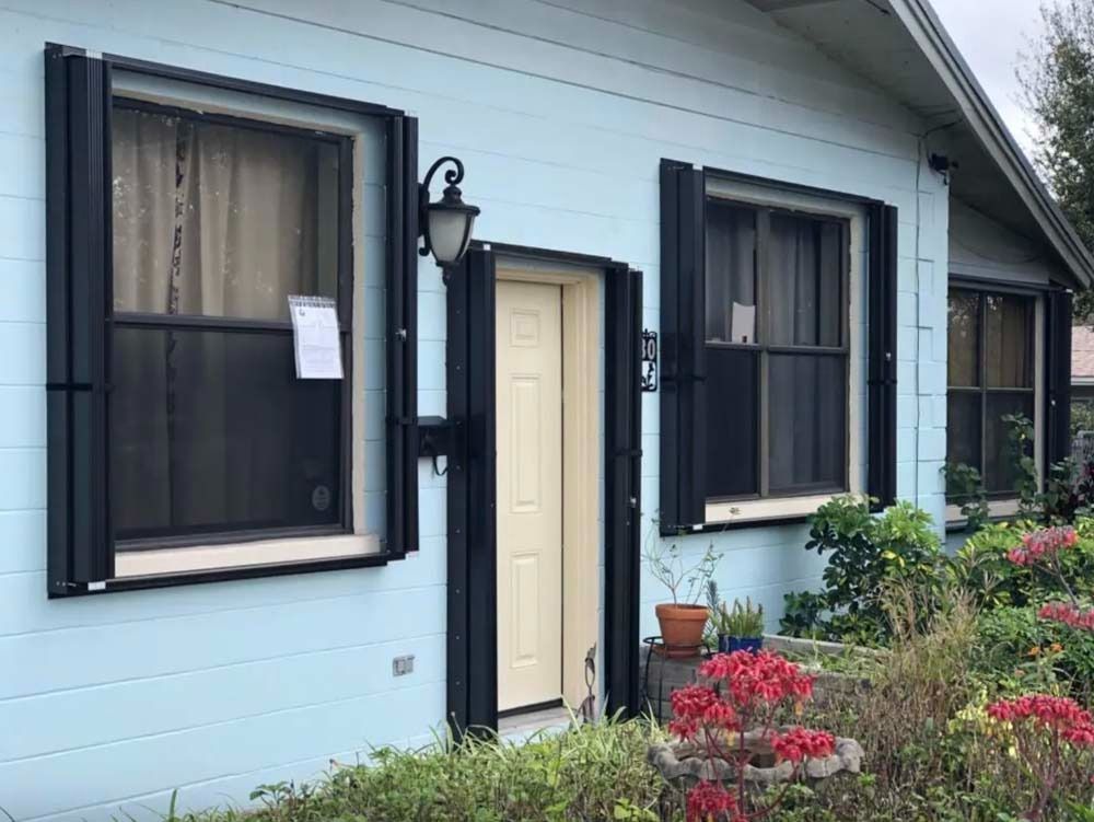 A light blue house exterior features a centered tan front door, dark trim, black hurricane shutters, and a front garden.