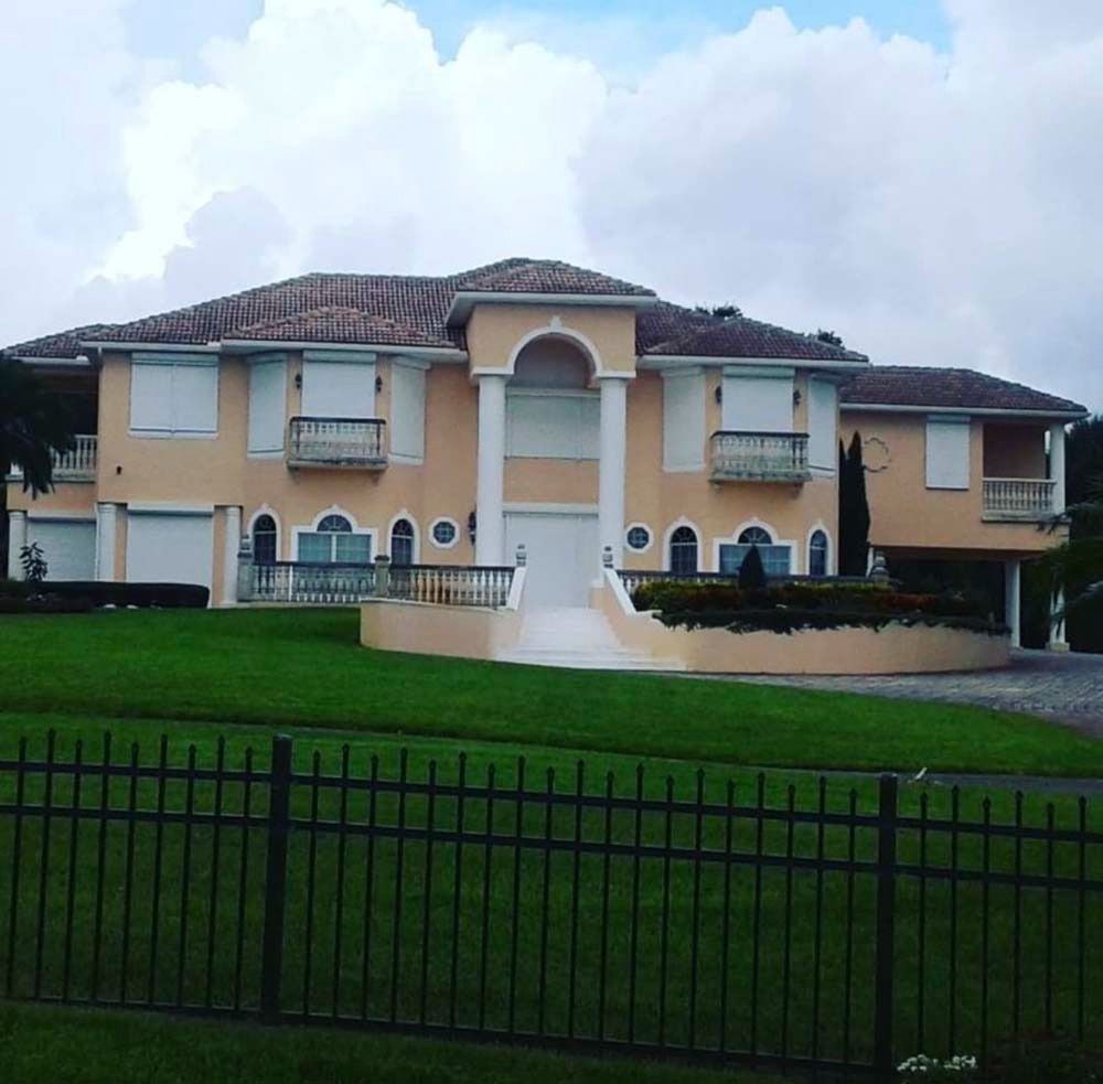 A two-story peach-colored house with a tiled roof, white columns, and hurricane shutters over the windows.