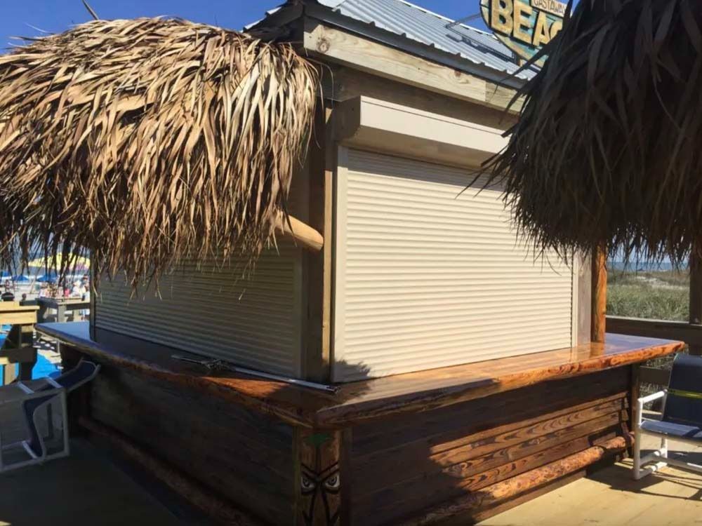 A closed outdoor tiki bar with a wooden counter, thatched roof, and tan rolling shutters on a sunny, sandy beach.