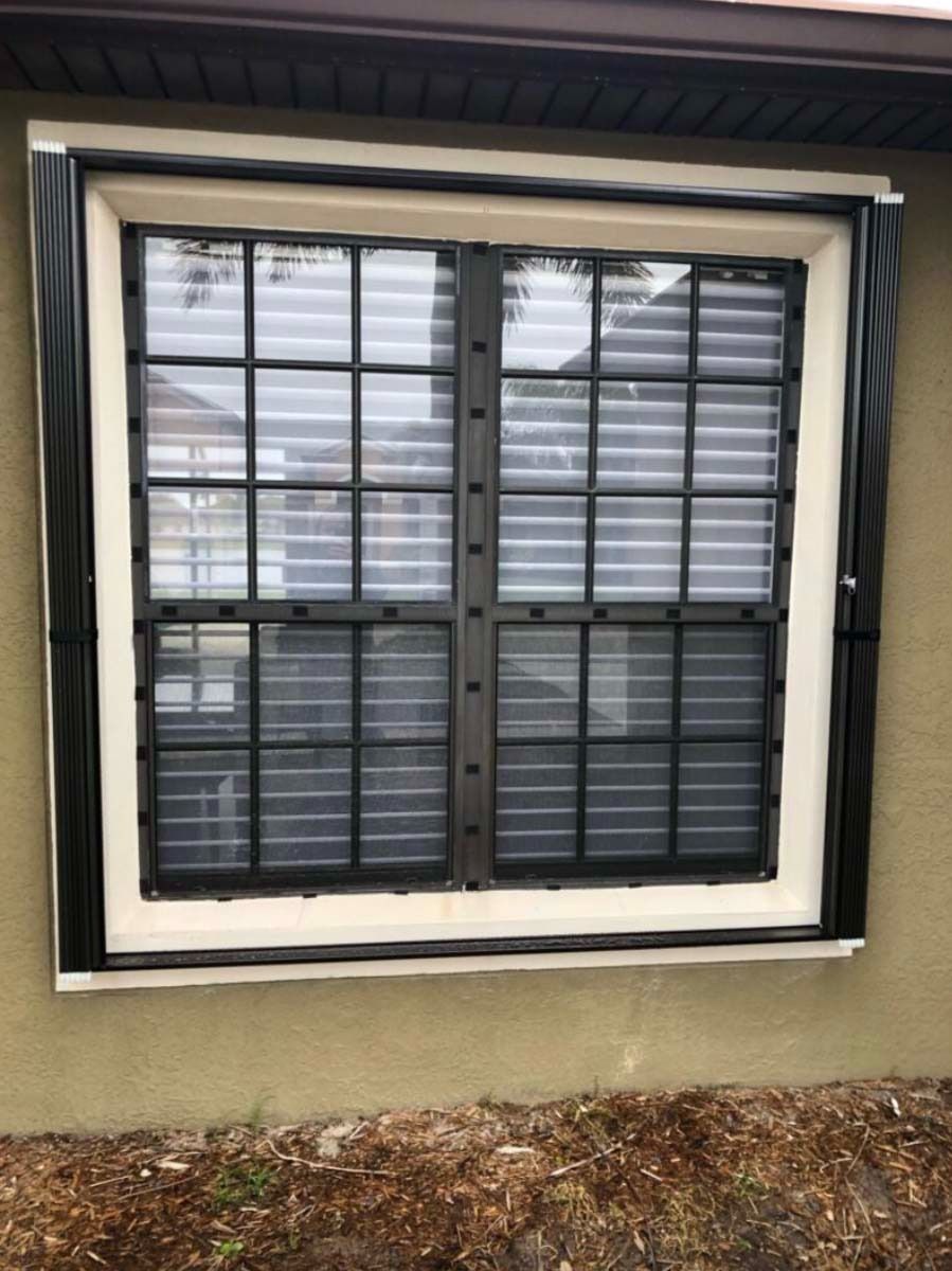 A black, frame-mounted window shutter system installed over a grid-patterned window on a light-colored stucco exterior.