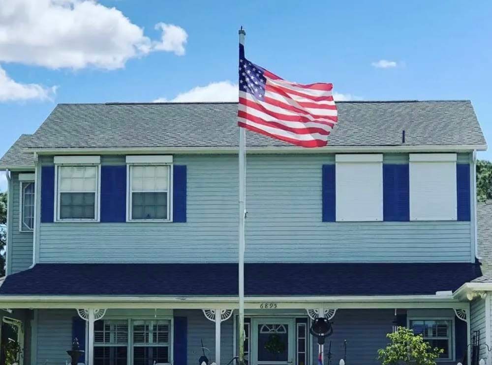 A light blue two-story house with blue shutters and a prominent American flag flying on a pole in the front yard.