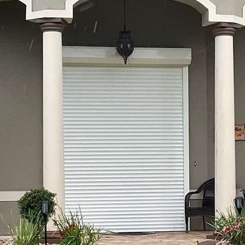 A white roller shutter covers the entryway of a home between two pillars, with a hanging lantern above and plants below.