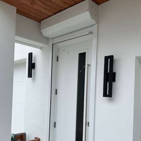 A white door featuring a vertical glass panel, centered under a roll-down shutter box, flanked by two black wall lamps.