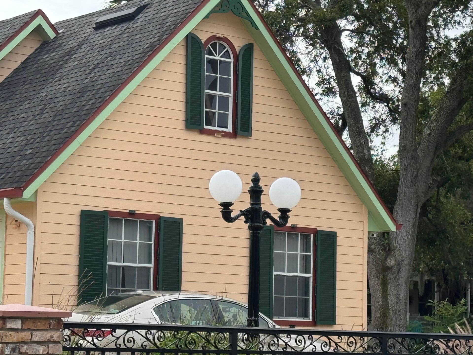 A light-yellow house with dark green shutters, a pointed roof, and a decorative double-globe street lamp in front.
