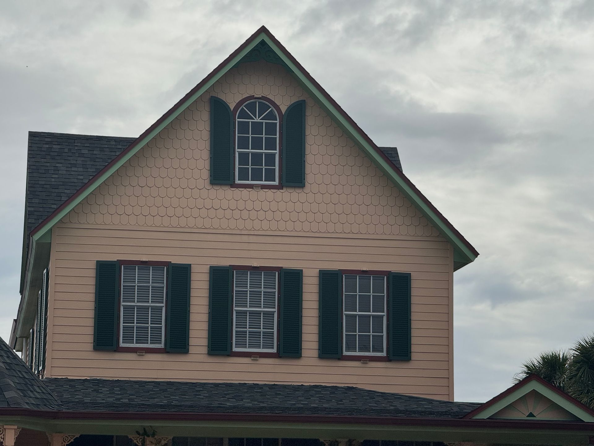 A salmon-colored house with a dark gabled roof, dark green shutters, and a patterned exterior under a cloudy sky.