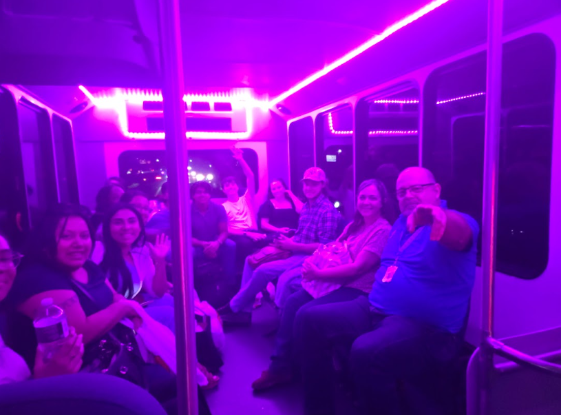 Passengers seated inside a neon-purple lit train car, one person pointing toward the camera.