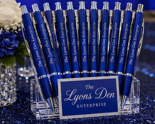 Blue “The Lyons Den” enterprise pens displayed in a clear holder on a glittery table