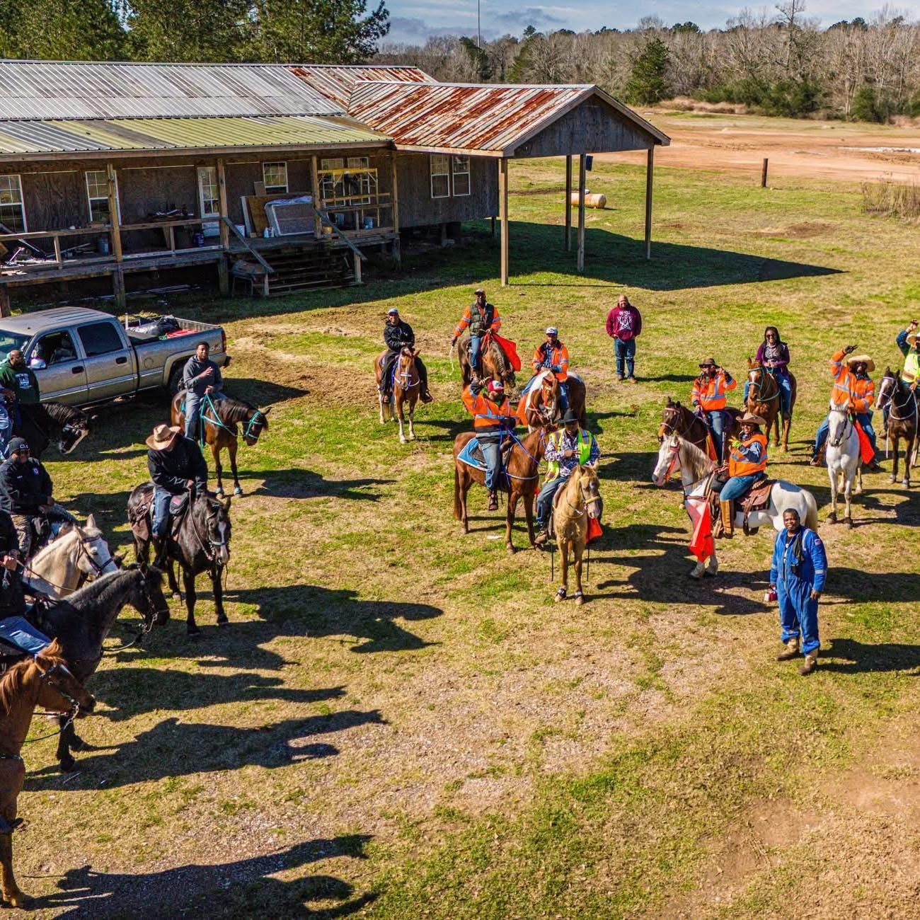 Group of horseback riders gathered in a grassy field beside a rustic barn under a clear sky
