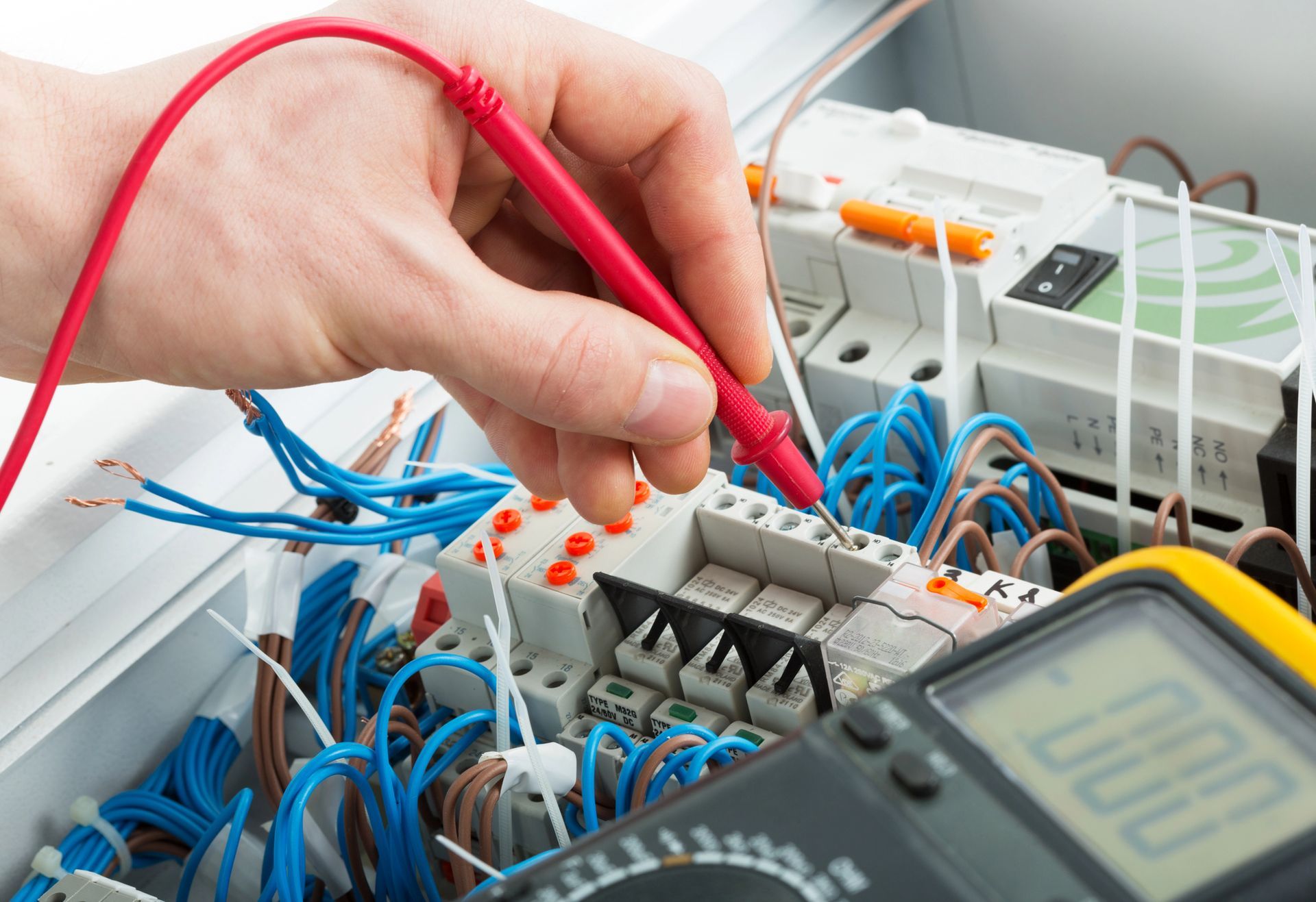 Una mano tiene la sonda rossa di un multimetro contro i morsetti elettrici all'interno di un quadro elettrico.