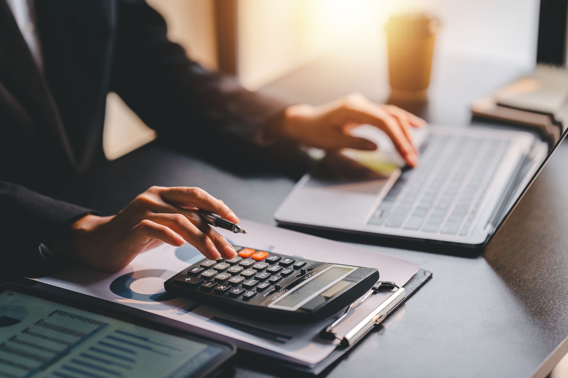 Person in a suit using a calculator and laptop at a desk with financial charts.