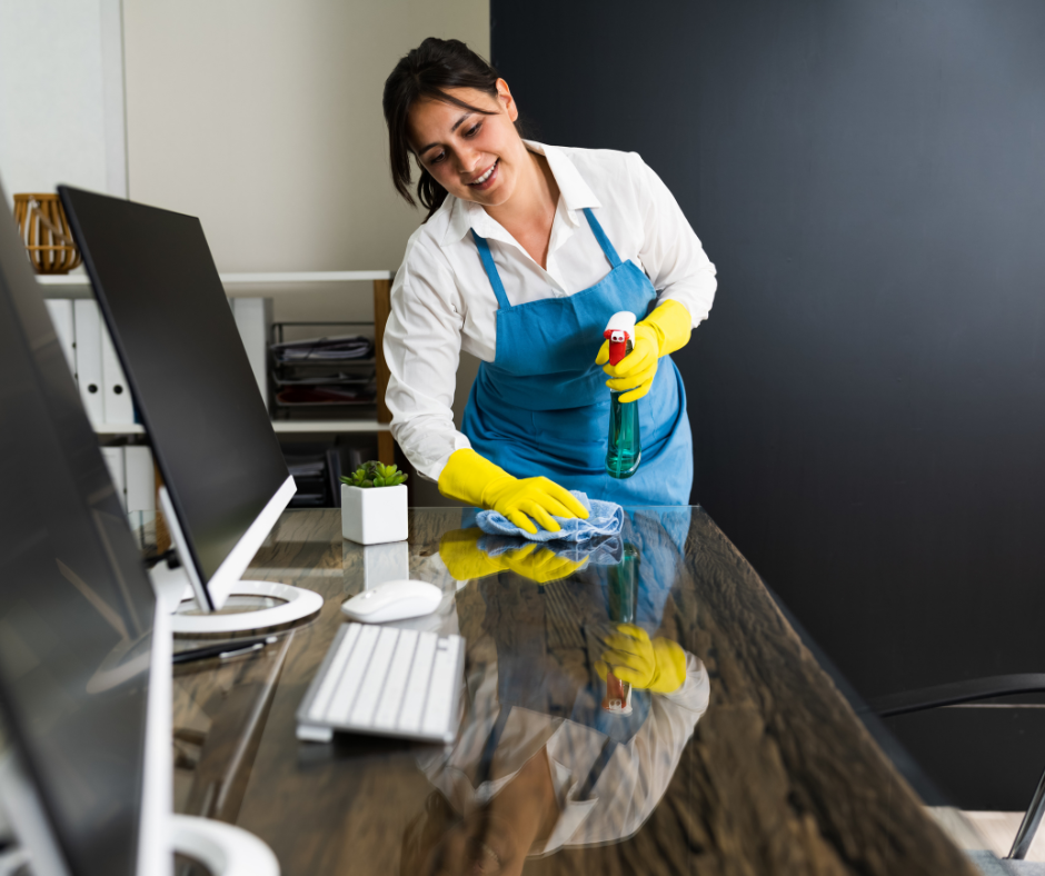 A person in a blue apron and yellow gloves wipes a dark office desk with a cleaning cloth and spray bottle.