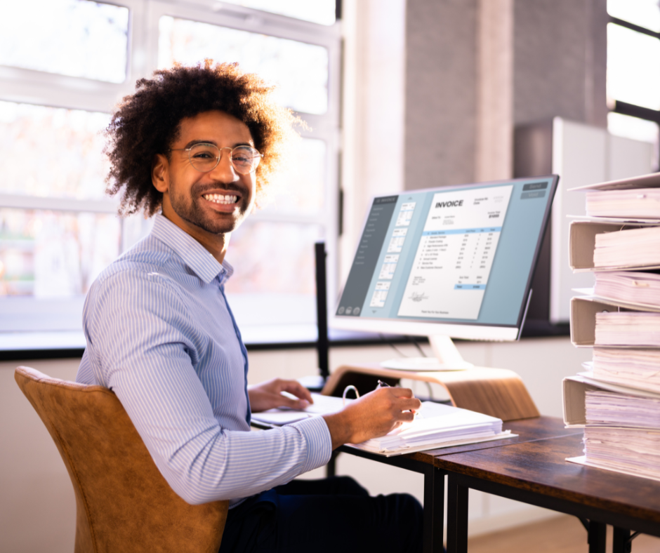A person in a light blue shirt smiles while writing at a desk with a computer displaying an invoice and stacks of binders.