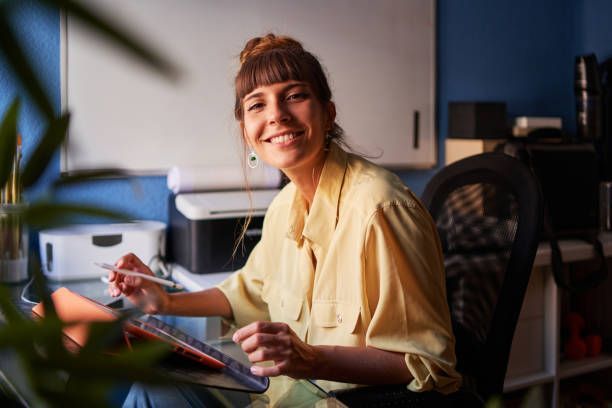 Woman in a yellow shirt smiles while working on a tablet at a desk in a home office.