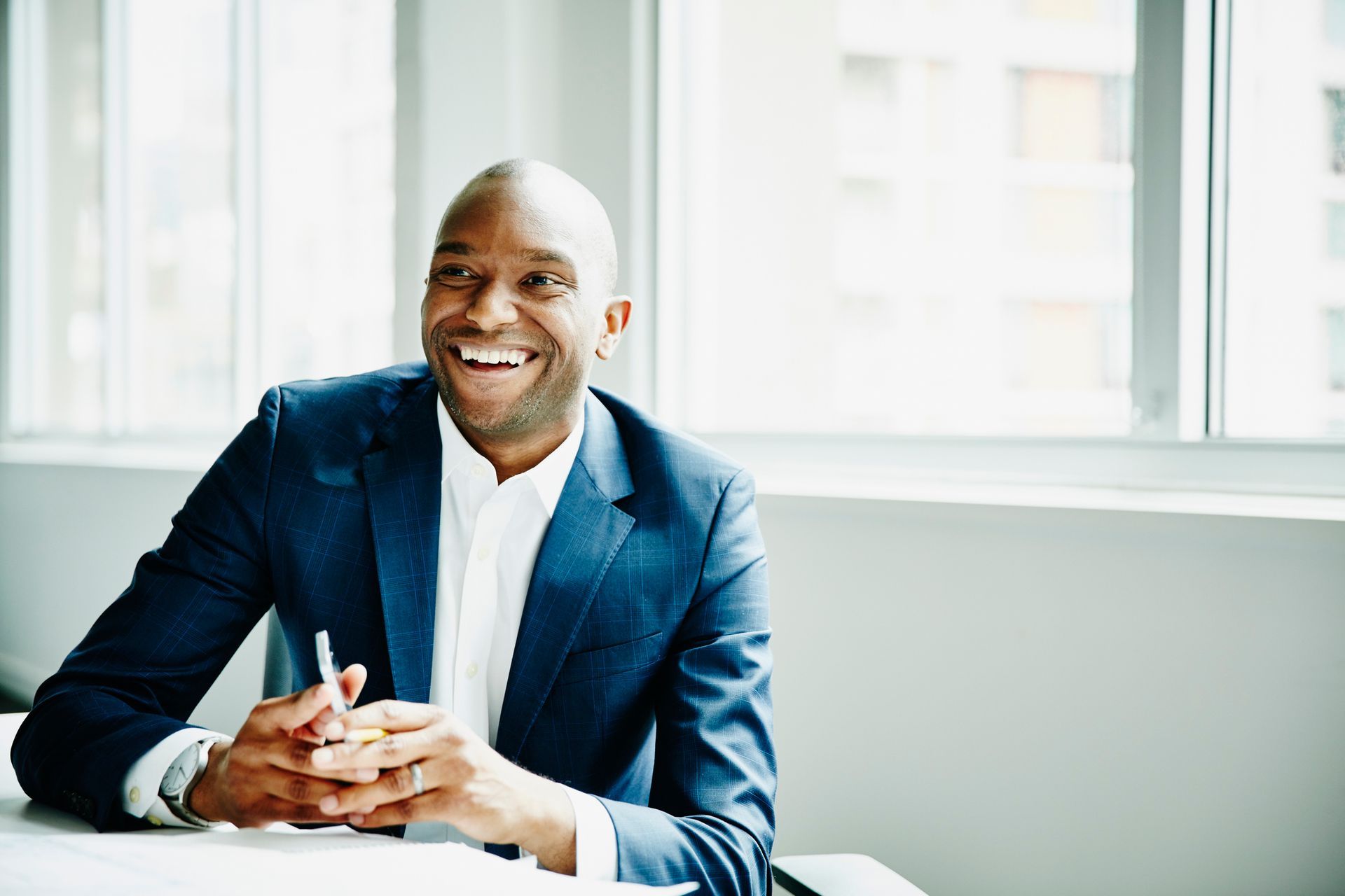 Man in blue suit, smiling, holding pen, seated at a desk near a window.