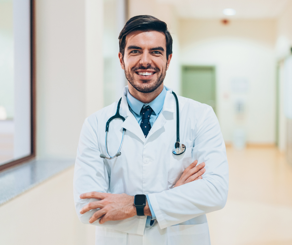 A smiling doctor in a white coat and stethoscope stands with arms crossed in a bright hospital hallway.