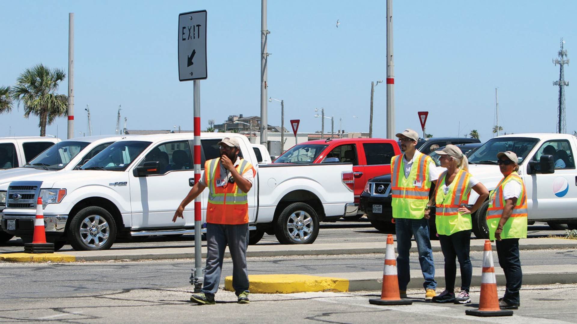 Workers in safety vests direct traffic near parked white pickup trucks on a sunny day.