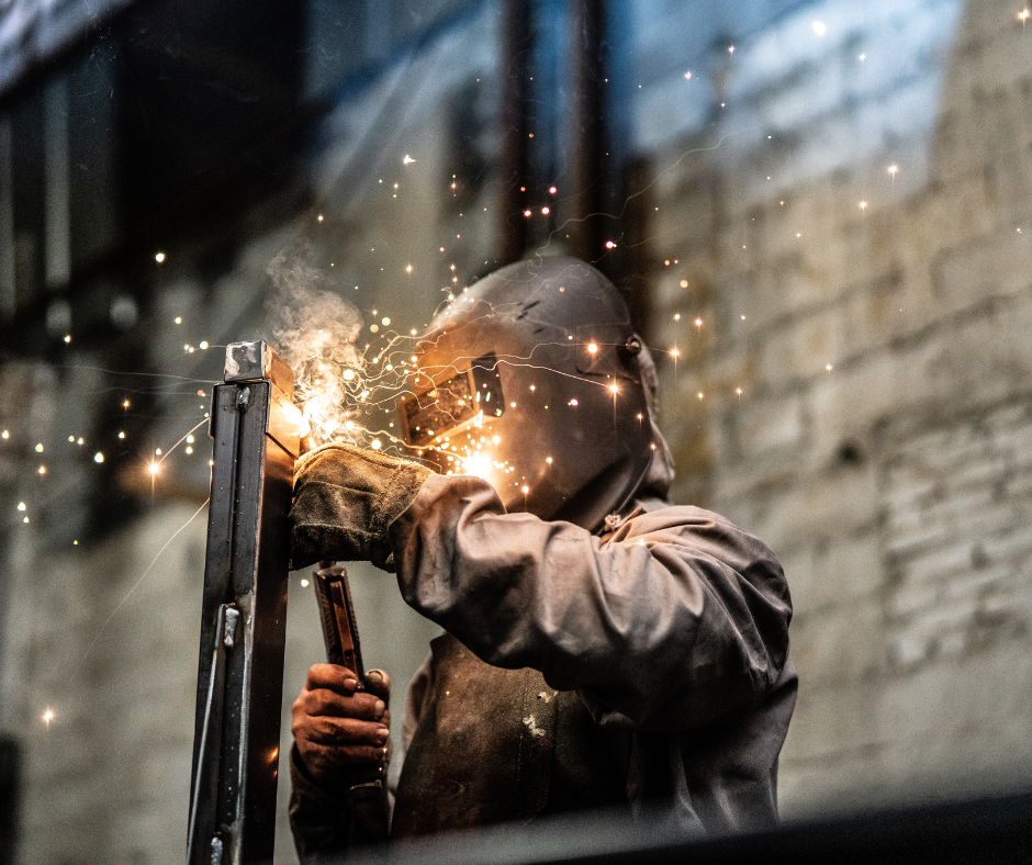 A welder in protective gear works on a metal beam, surrounded by a shower of bright orange sparks in an industrial space.