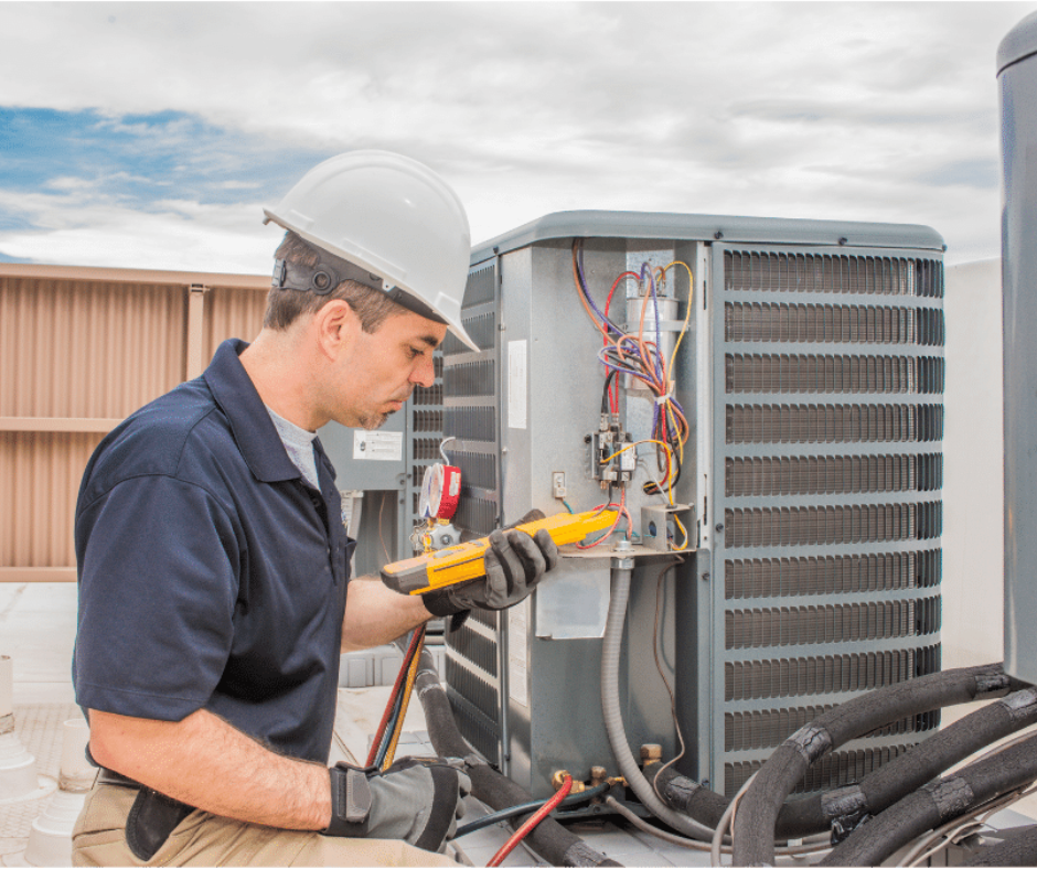 A technician in a white hard hat and work clothes uses a yellow multimeter to test an outdoor HVAC unit's wiring.
