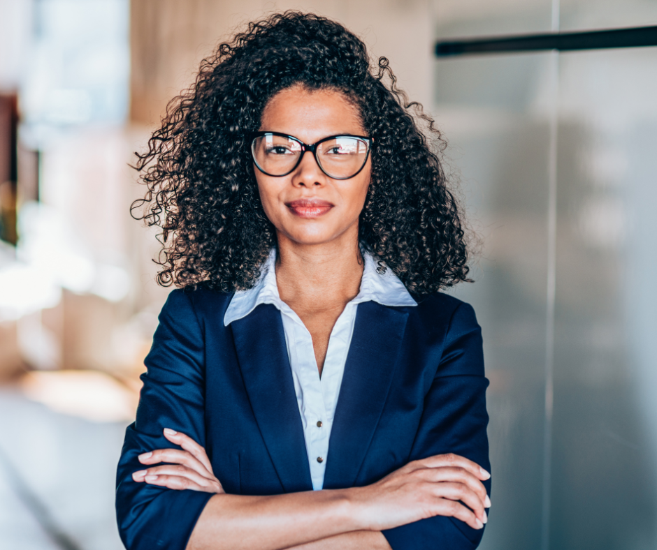 A professional woman with curly hair and glasses wears a blue blazer and white shirt, standing with arms crossed.