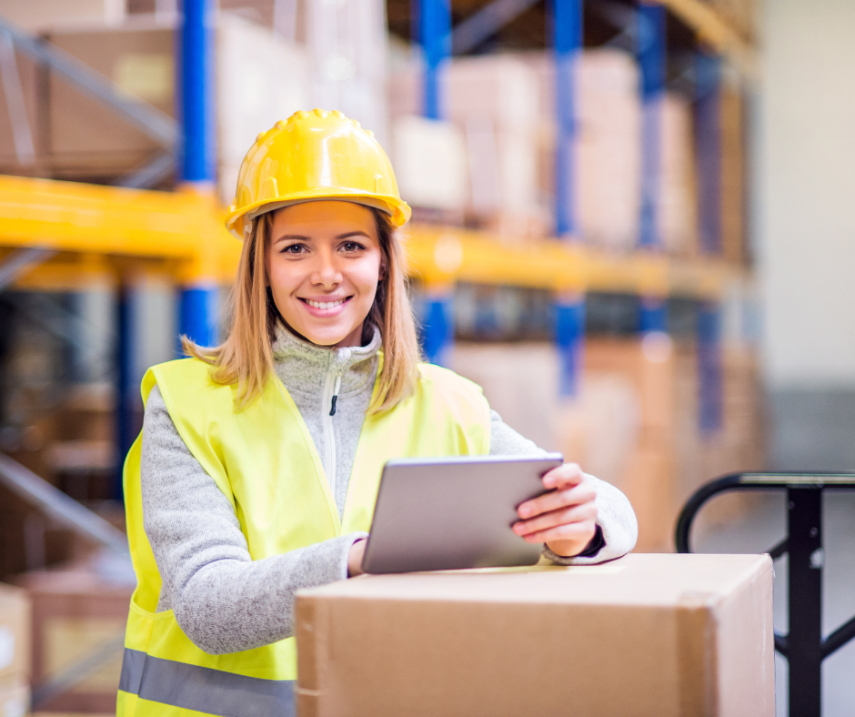 A smiling warehouse worker in a yellow hard hat and safety vest holds a tablet while leaning on a cardboard box.