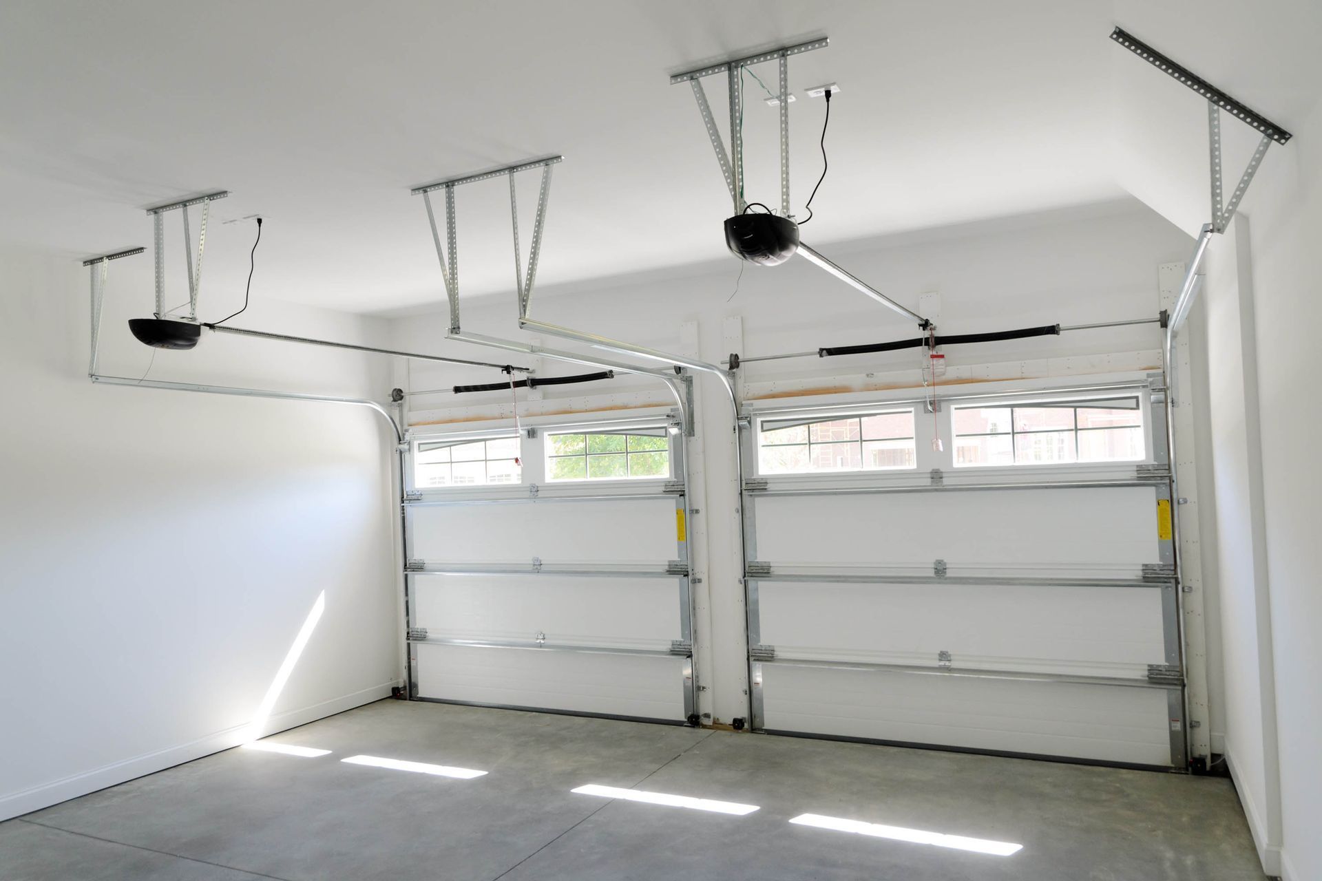 A garage interior with two open white doors, showing ceiling-mounted garage door openers and concrete floor.