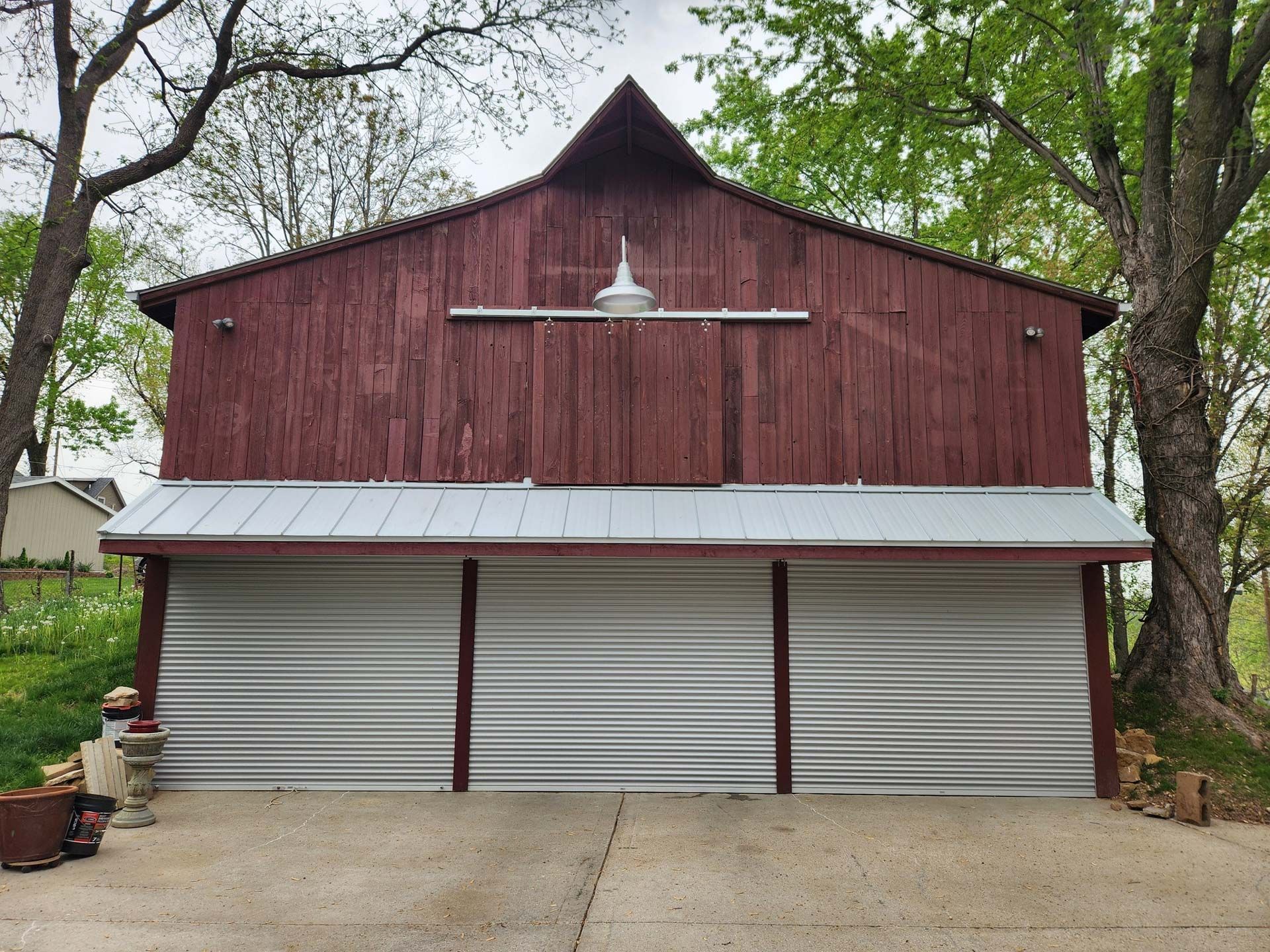 Red wooden barn with corrugated metal doors, concrete driveway, and surrounding trees.
