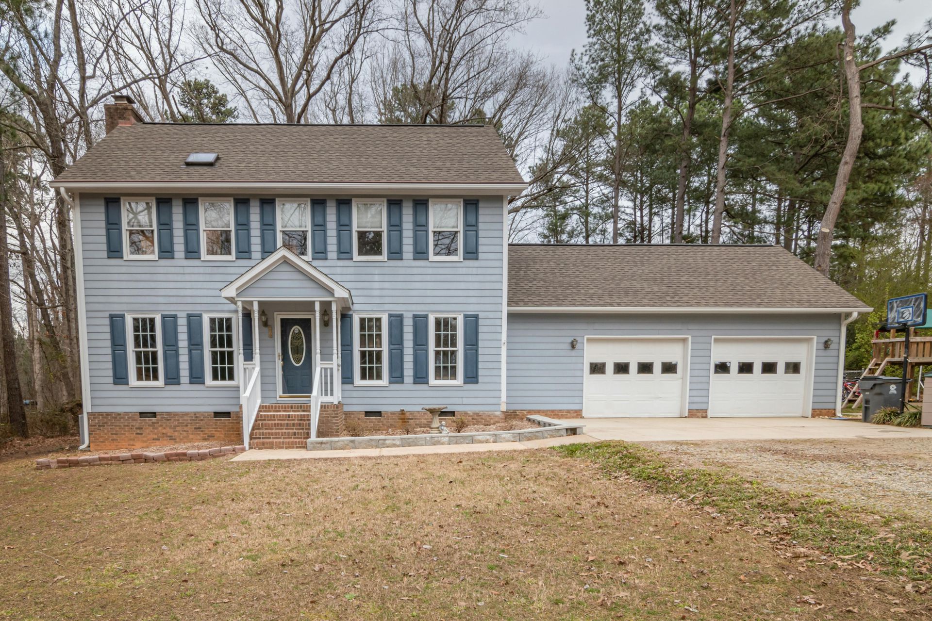 Two-story blue house with white trim, shutters, and two-car garage. Brown roof and bare trees surround it.
