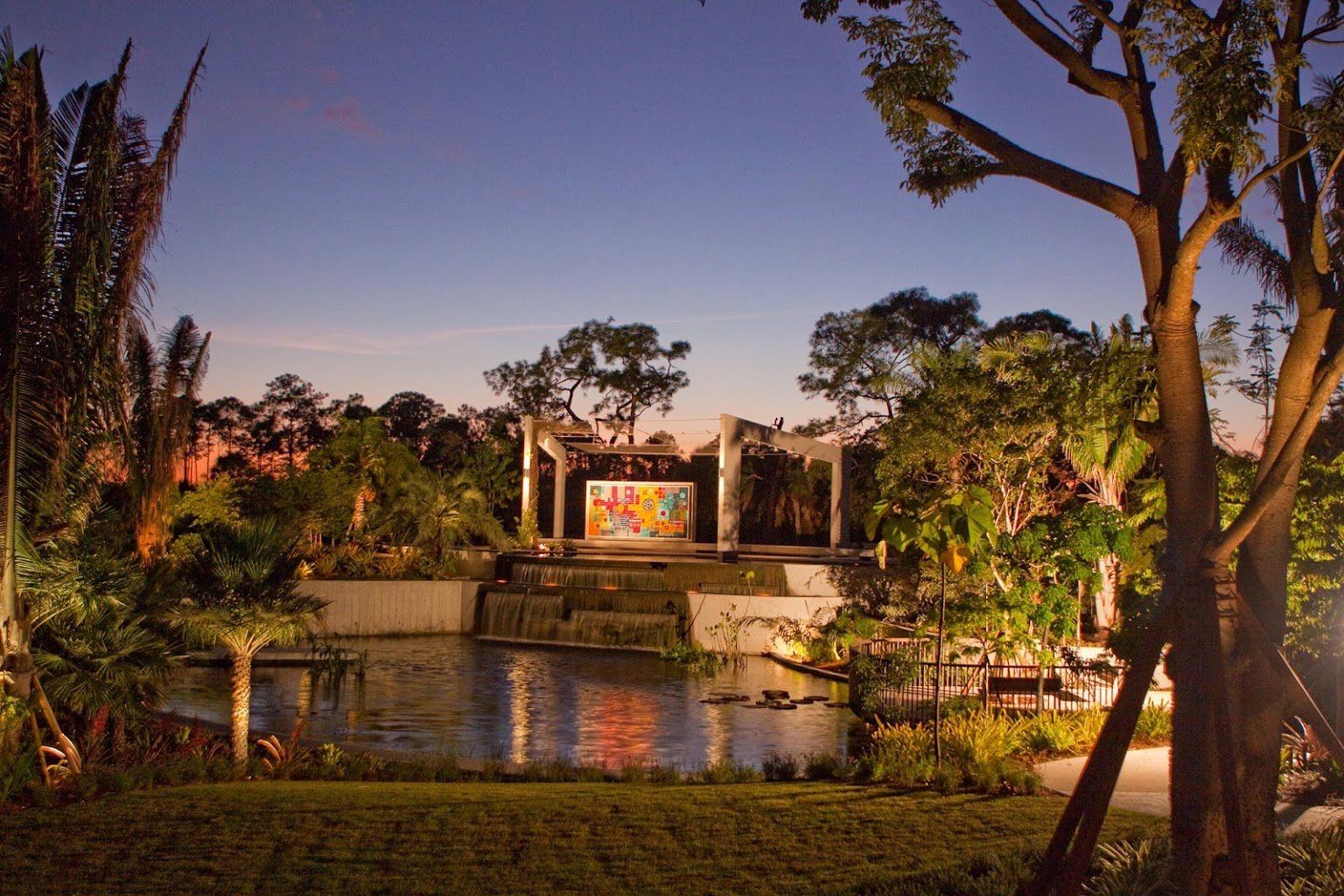 Sunset view of a landscaped garden with a reflective pond, trees, and a lit pavilion in the distance
