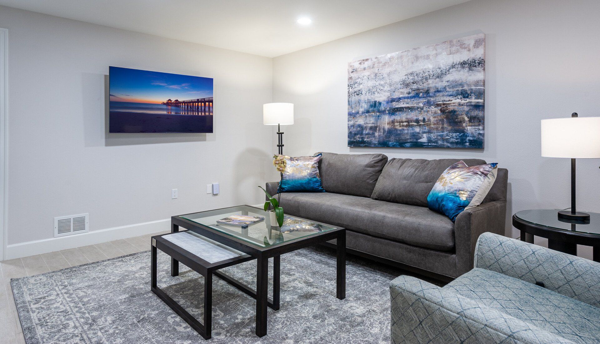 Modern living room with gray sofa, glass coffee table, patterned rug, and wall art in a bright space