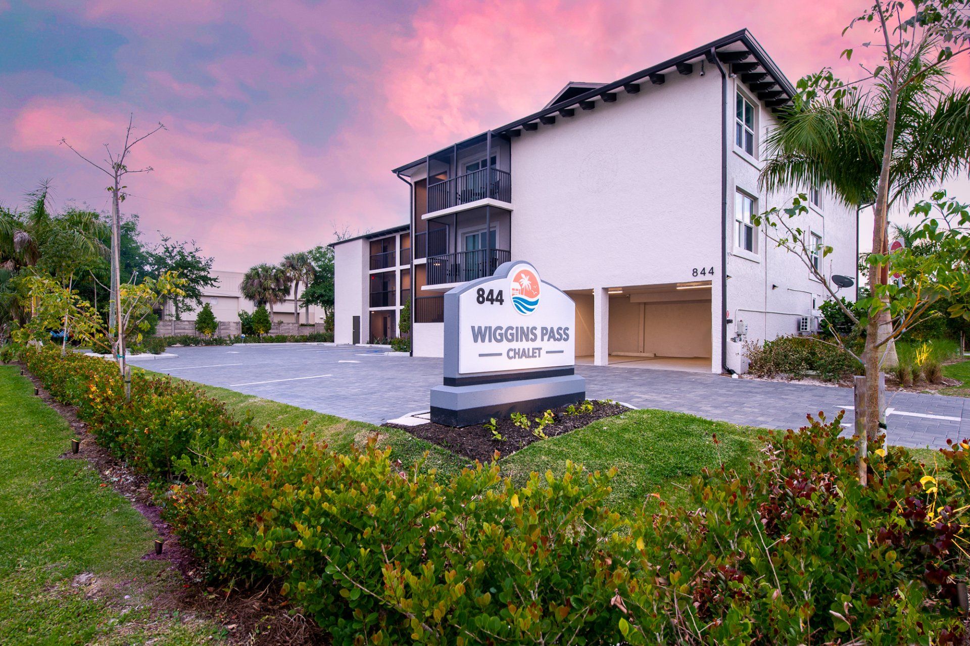 Modern apartment building at dusk with a “Welcome to” sign, landscaped shrubs, and pink sky