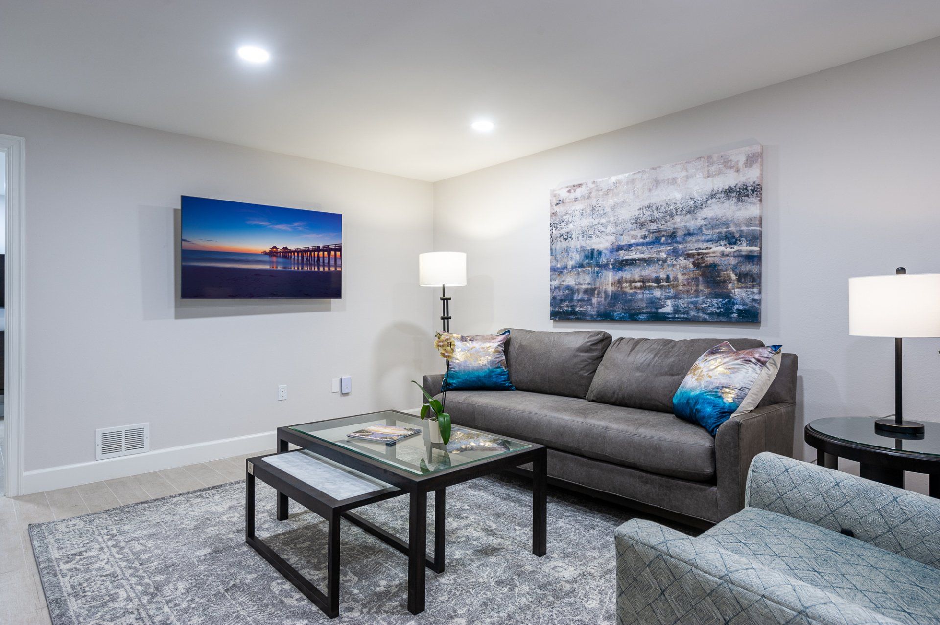 Modern living room with gray sofa, blue accents, glass coffee table, and framed wall art