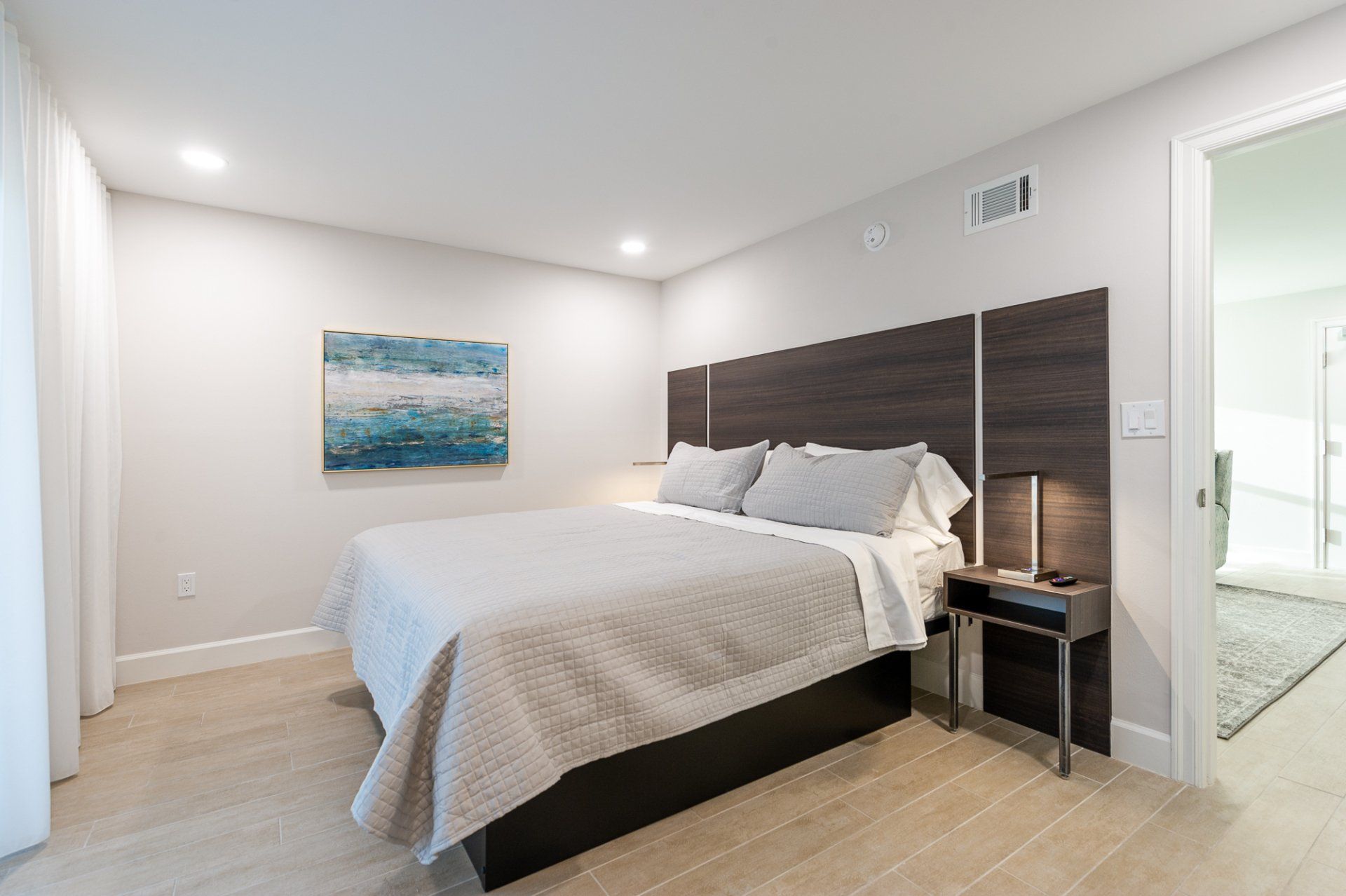Bright modern bedroom with a made bed, dark wood headboard, and white walls.