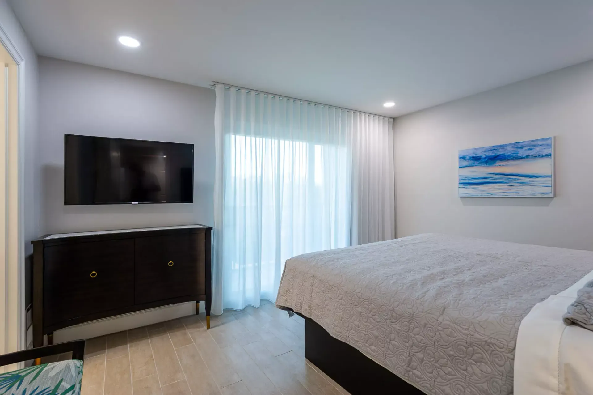 Bright bedroom with a large bed, wall-mounted TV, dresser, sliding glass door, and coastal painting