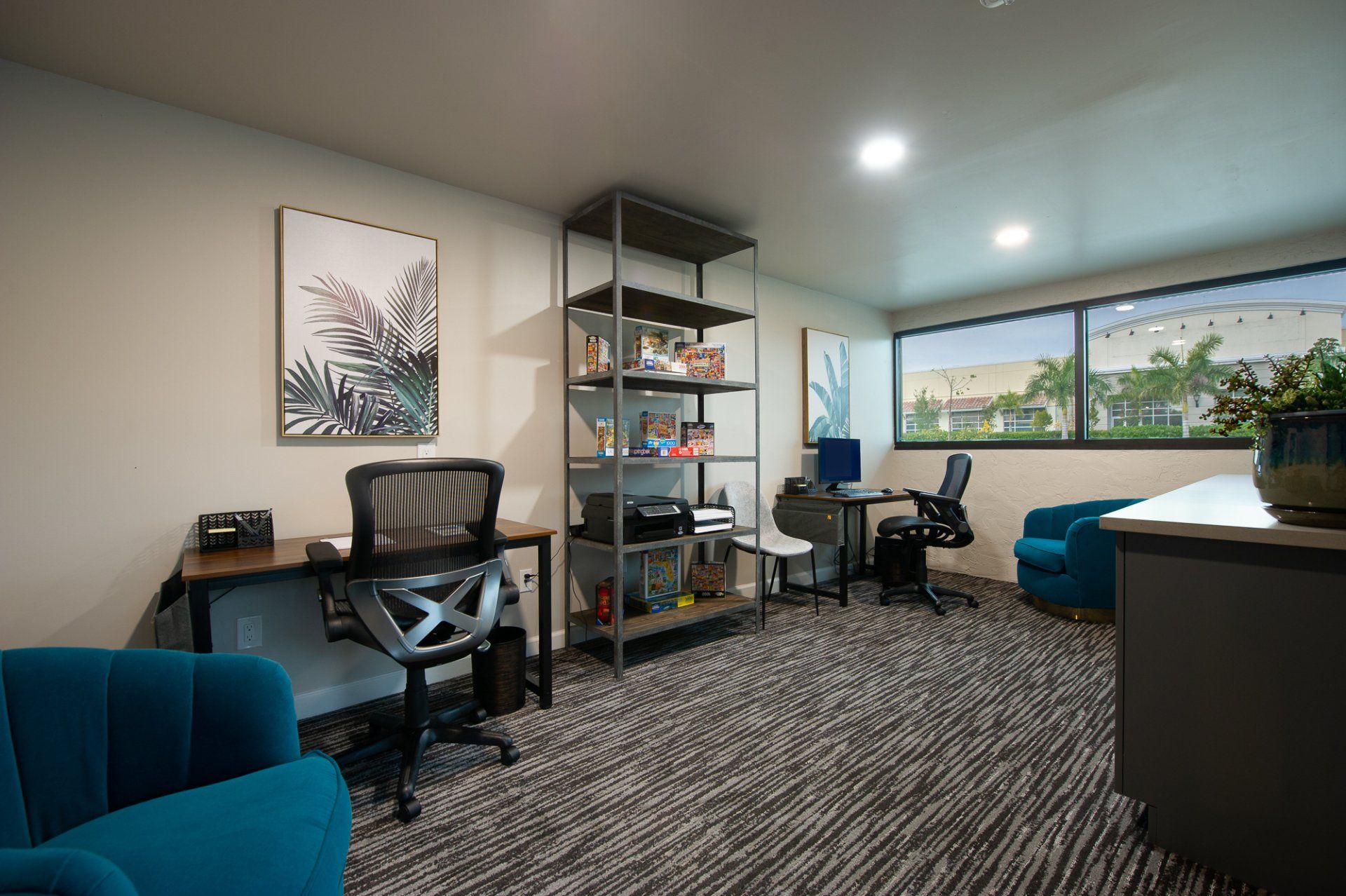 Modern office room with desks, chairs, shelves, and large windows overlooking greenery.