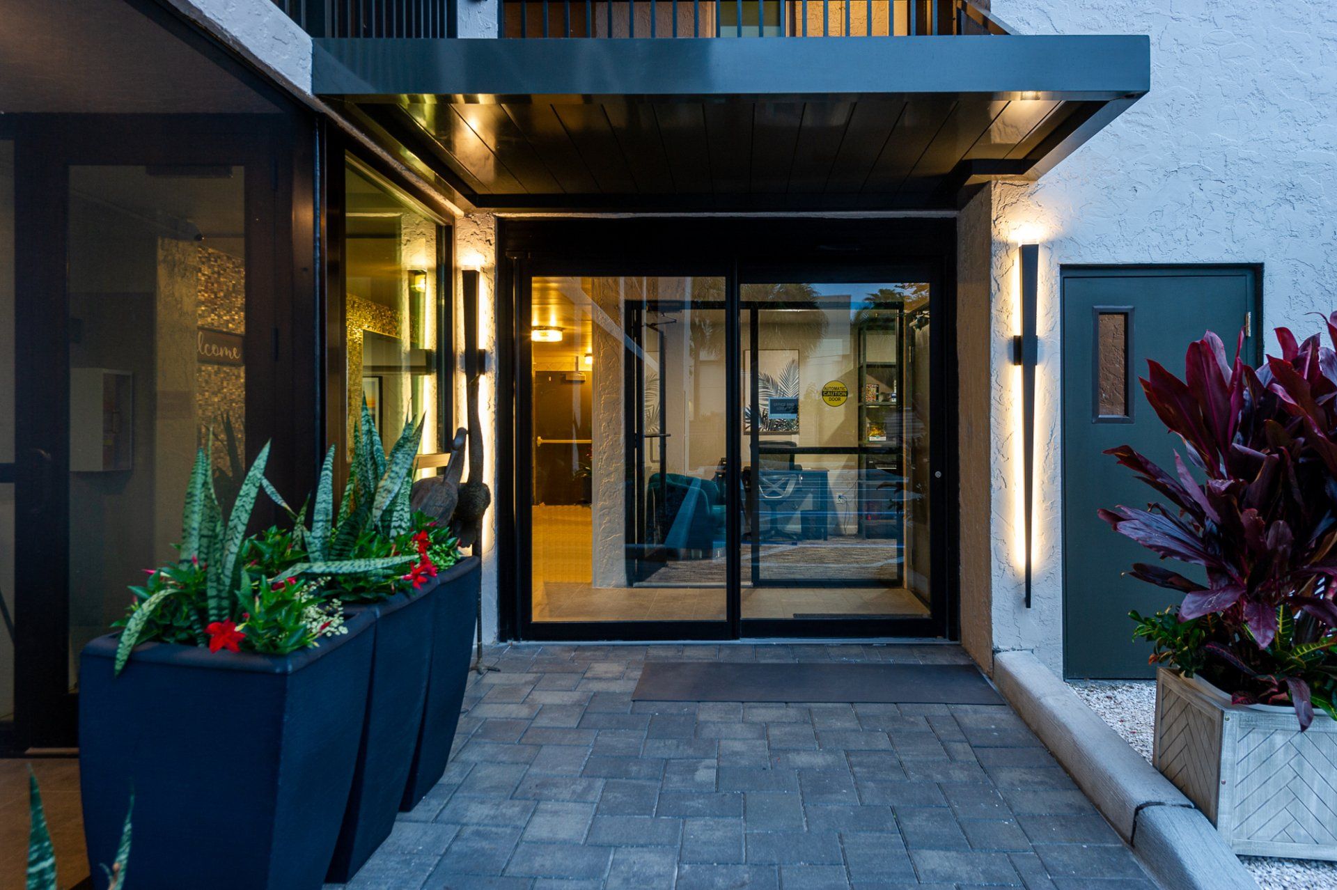 Modern building entrance with glass doors, dark awning, potted plants, and stone walkway at dusk