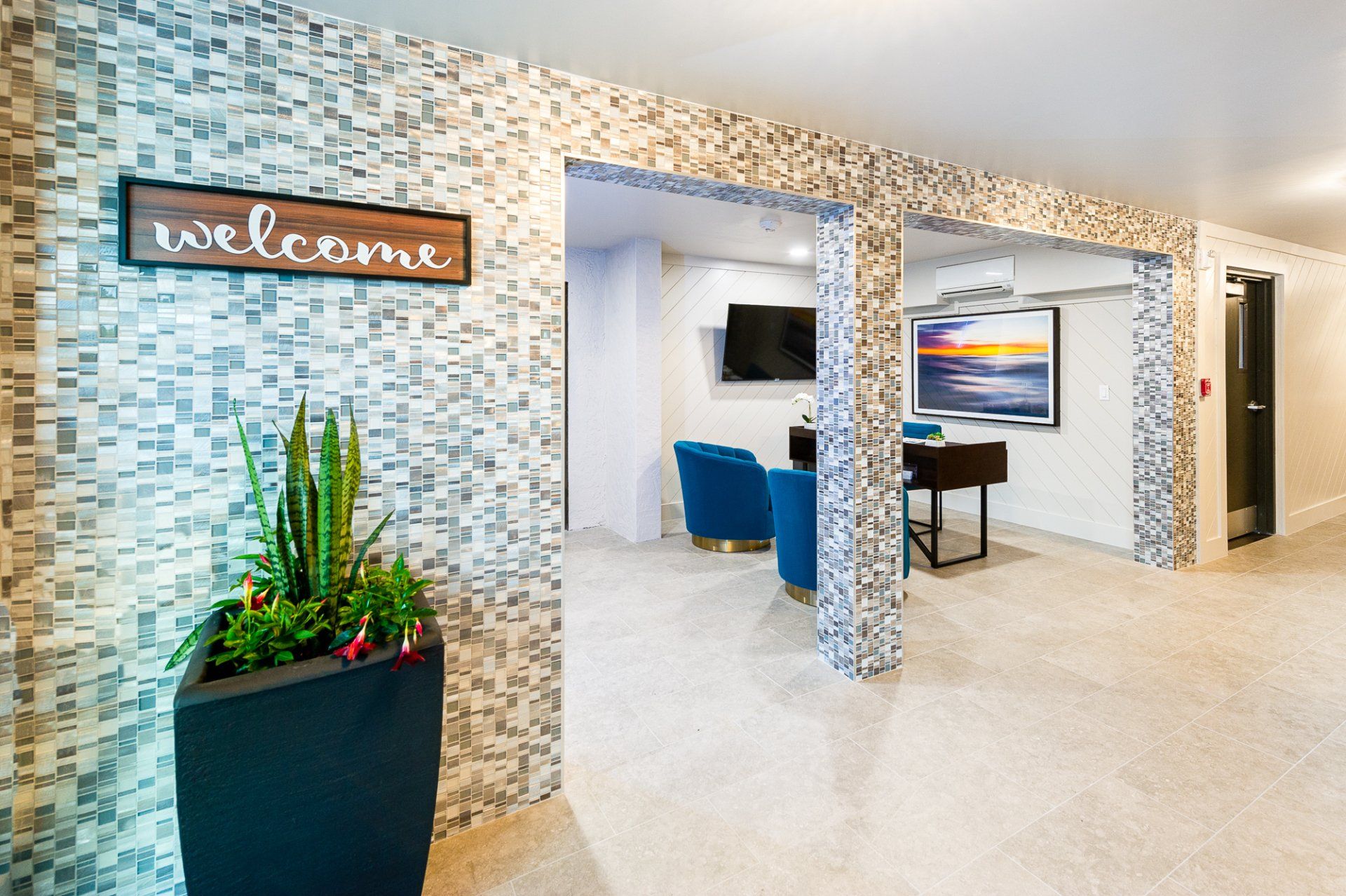 Modern office lobby with mosaic walls, a “Welcome” sign, blue chairs, and a large plant.