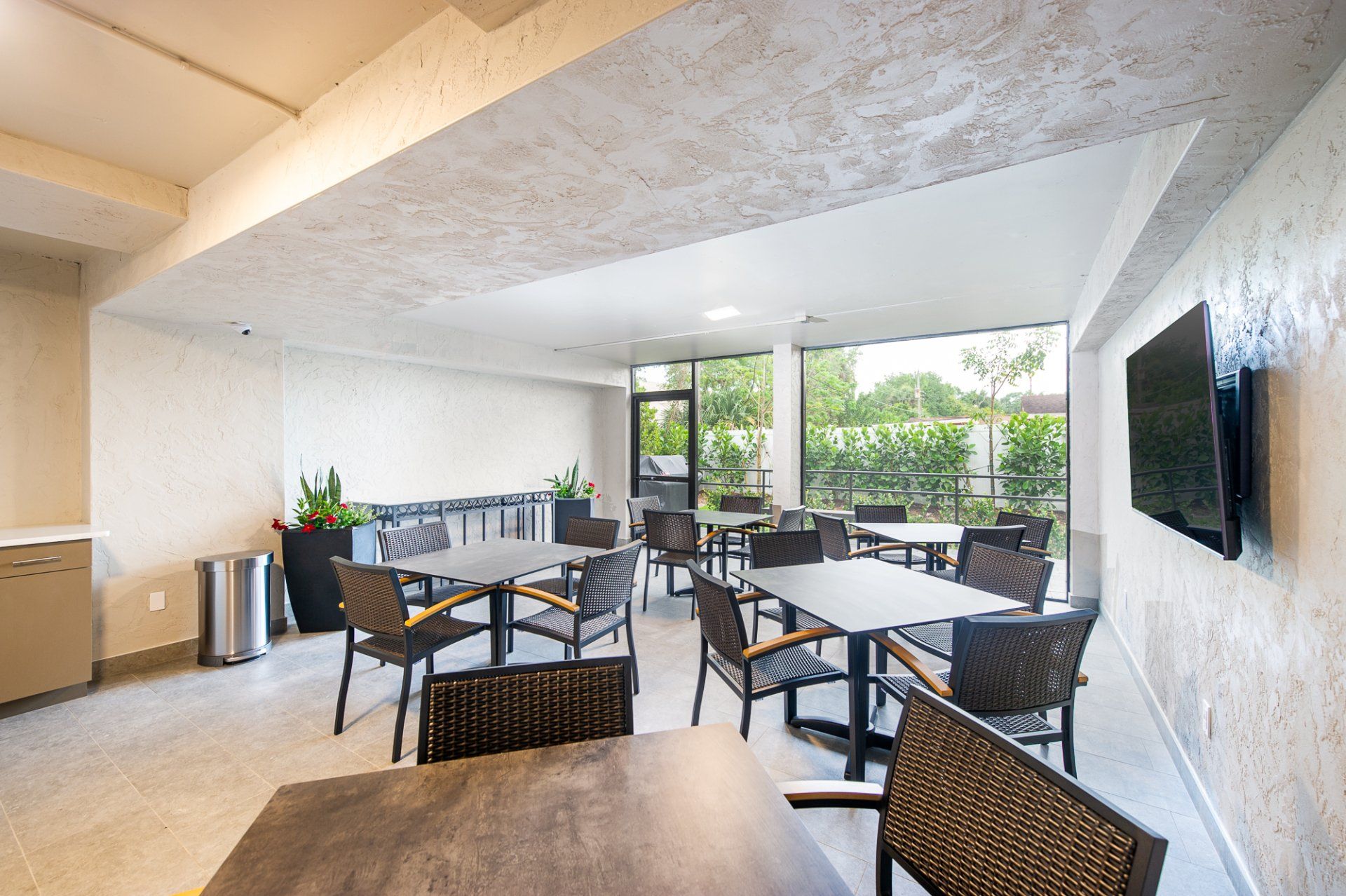 Bright lounge with tables and chairs, large windows, and a wall-mounted TV overlooking greenery