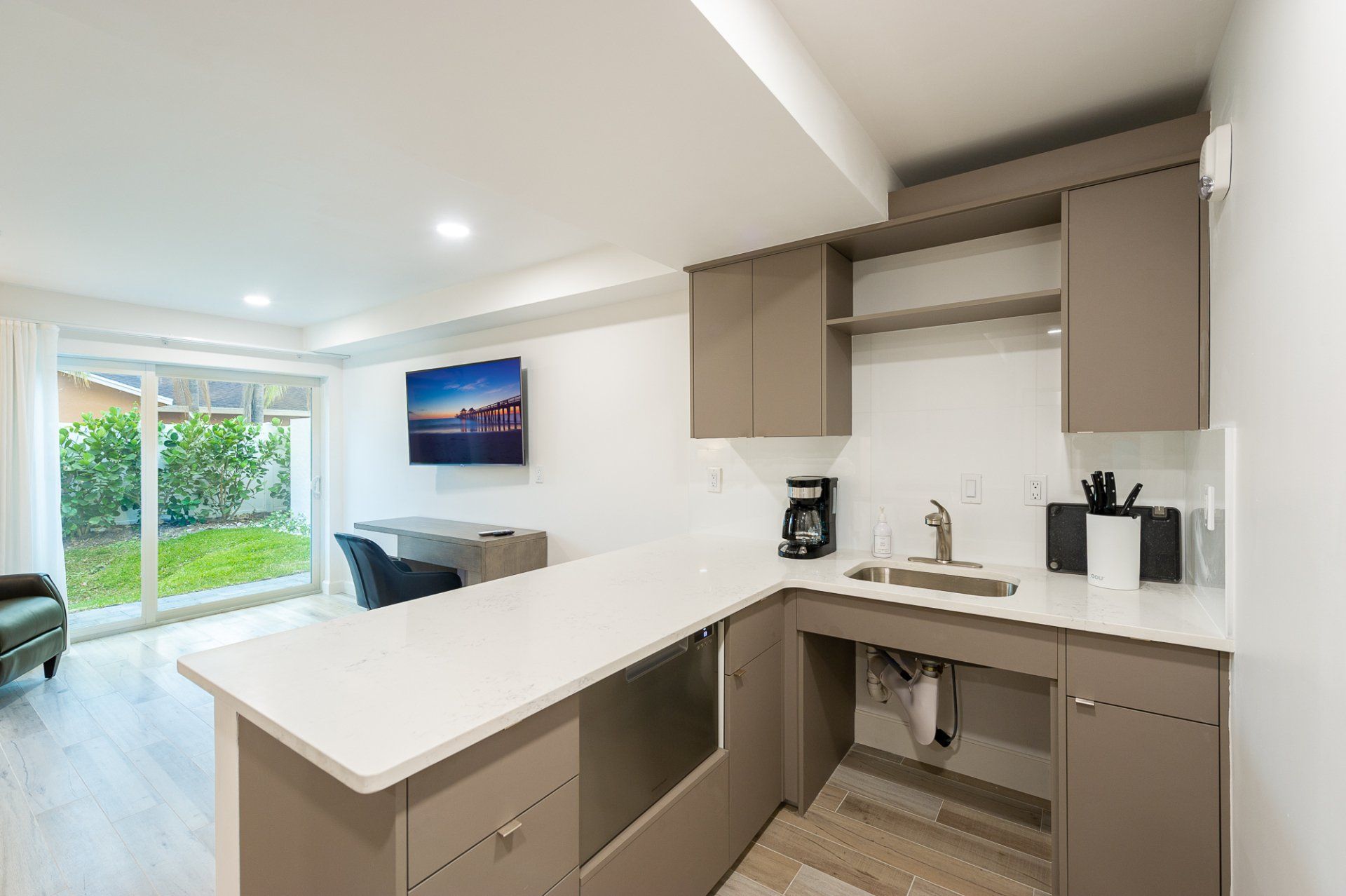 Modern kitchen with white countertops, beige cabinets, and a view into a bright living area with sliding glass doors.