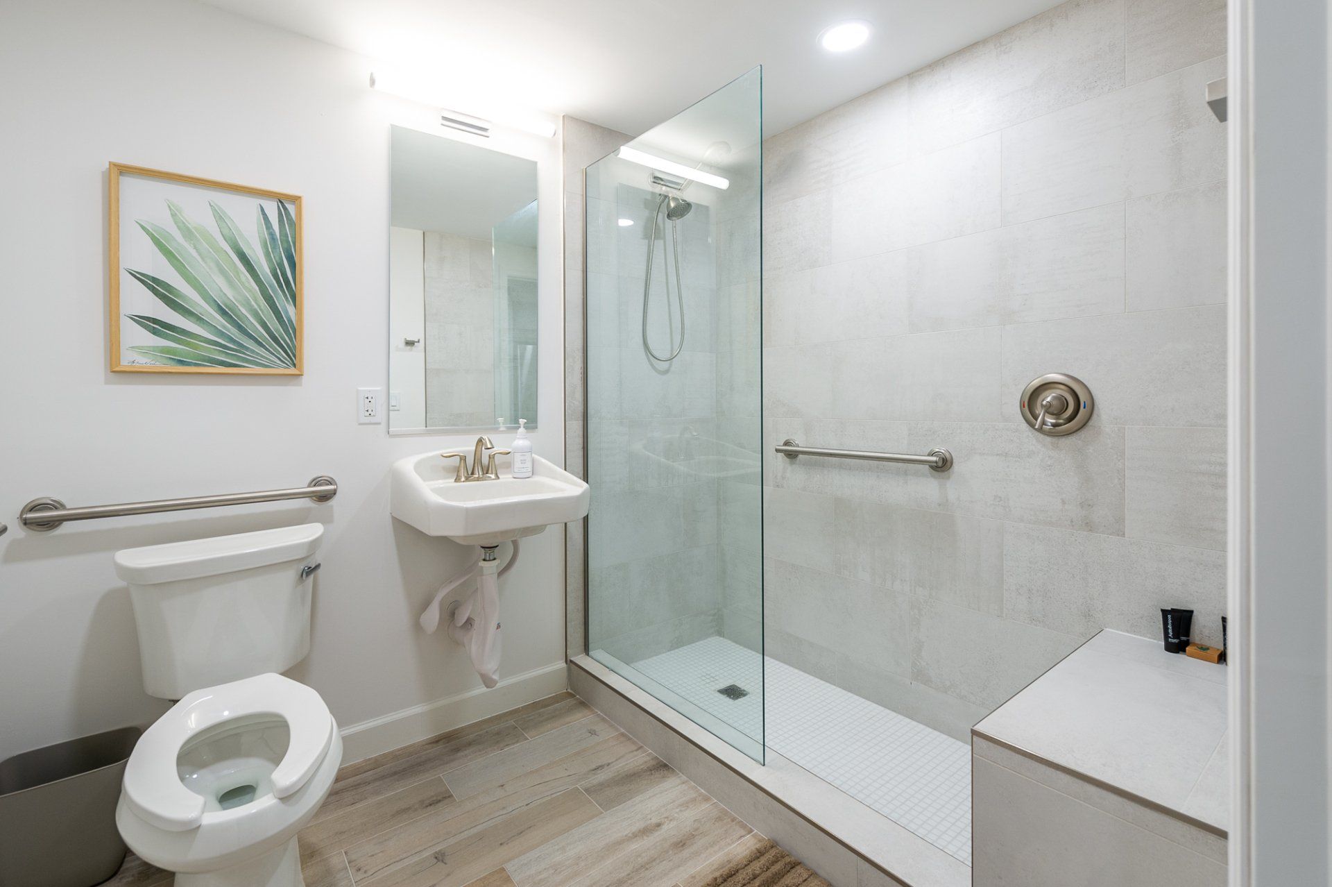 Bright accessible bathroom with toilet, sink, glass shower, and grab bars on white tile.
