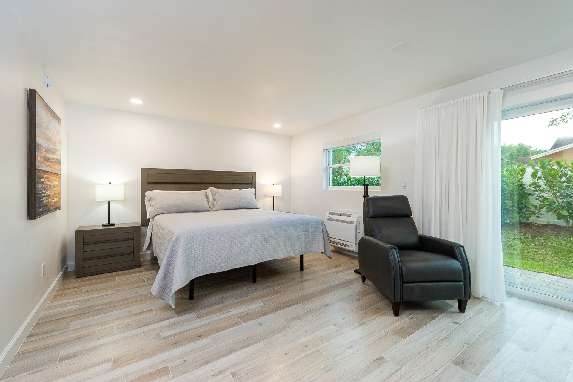 Bright bedroom with a king bed, nightstands, black recliner, and large window overlooking a yard