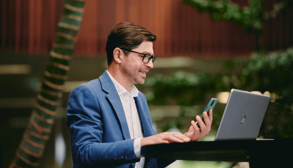 Man in blue blazer smiling at a phone beside an open laptop outdoors