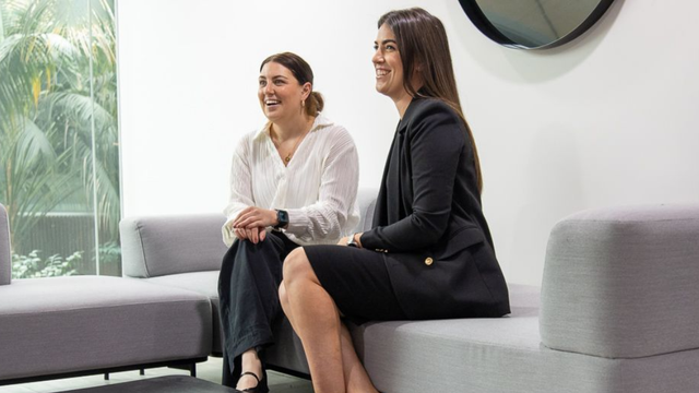 Two women in business attire sit on a gray couch, smiling and talking in a bright modern office lounge.