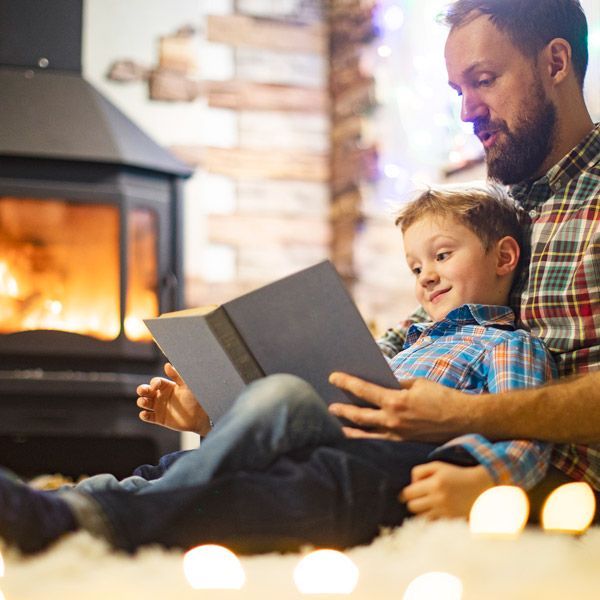 Adult reading a book with a child beside a lit fireplace in a cozy living room